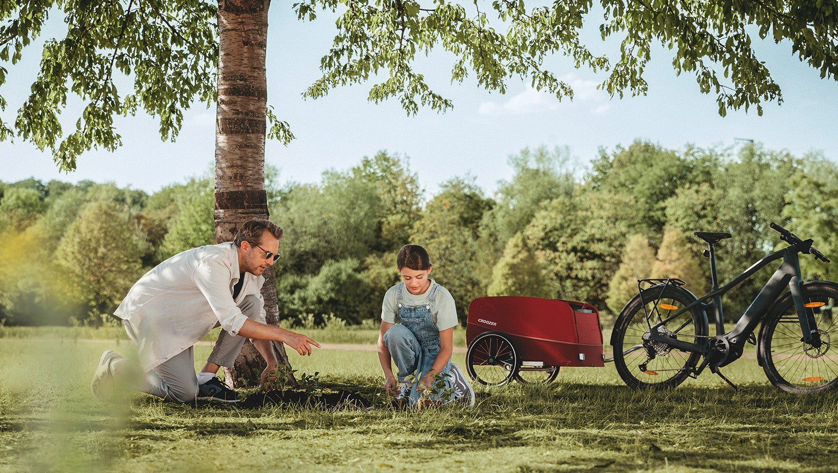 Father and daughter planting tree together in park with red Croozer cargo bike trailer and e-bike under tree
